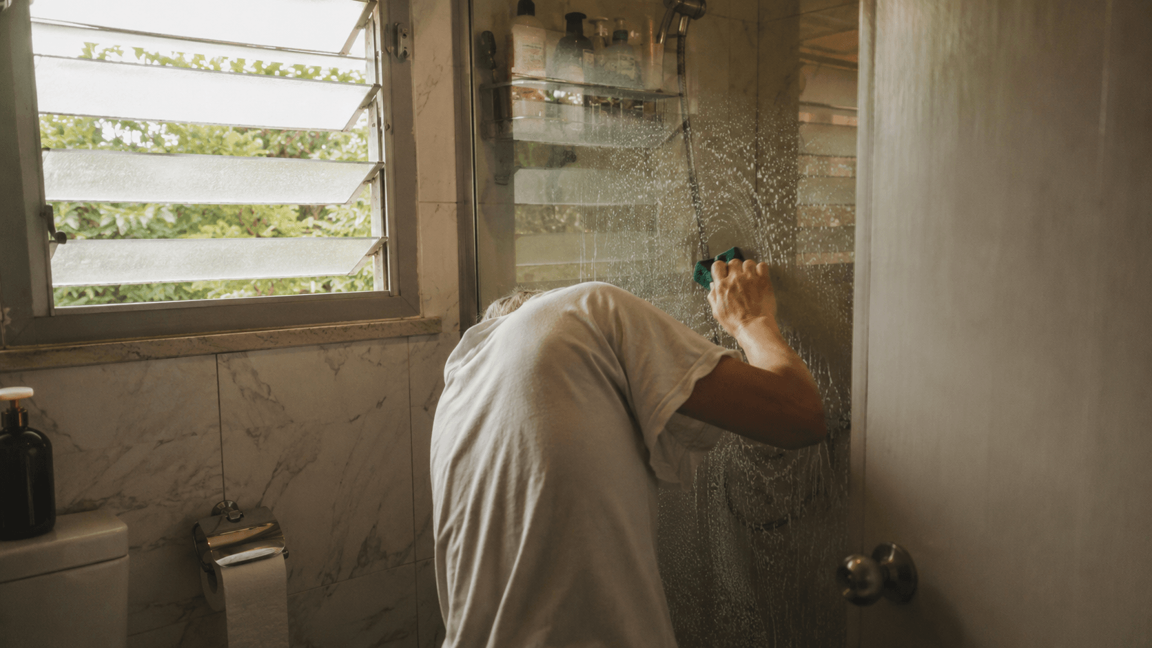 Quiet Saturday-morning moment of someone scrubbing a stained shower glass — the universal frustration that started everything