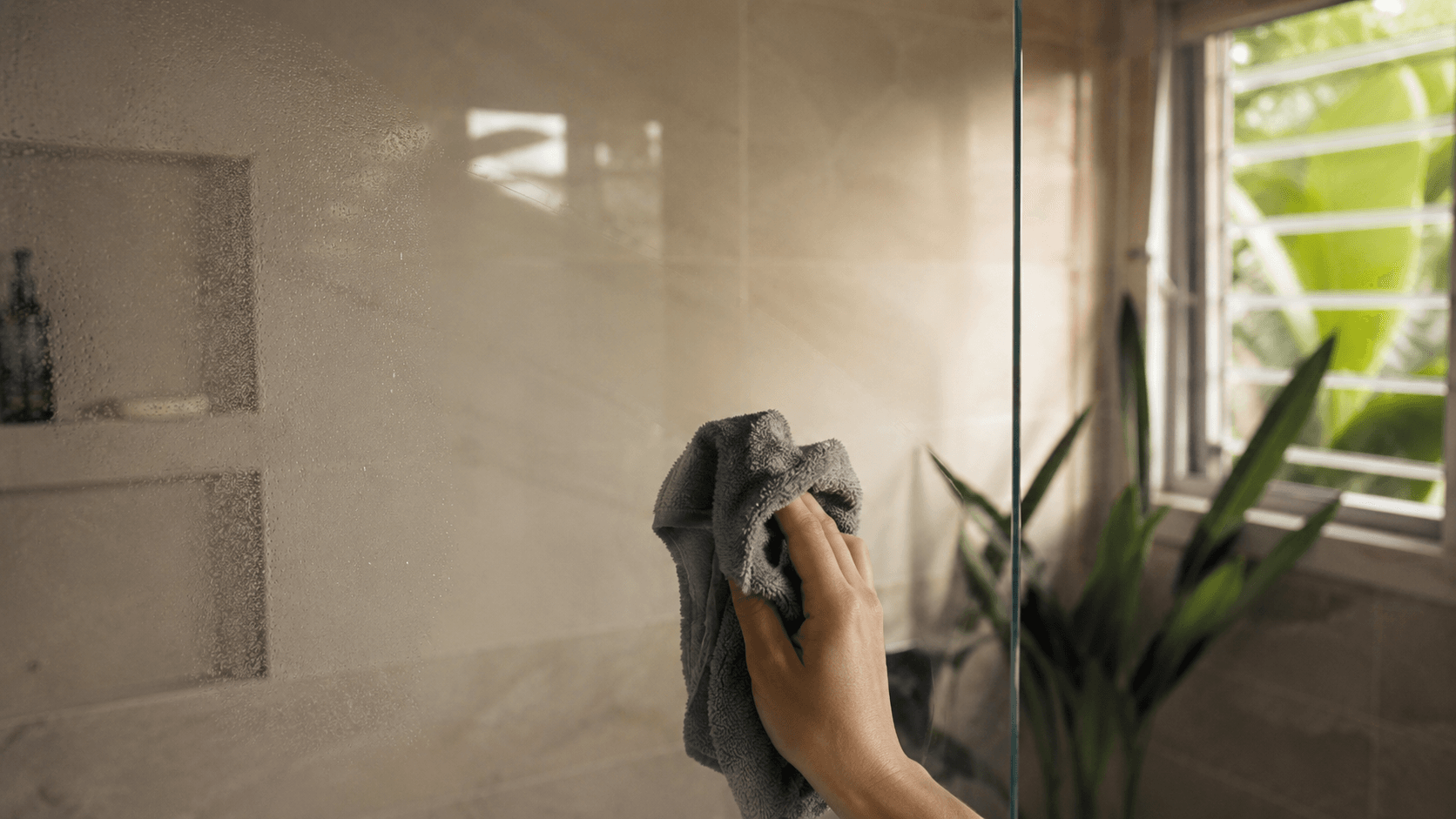 A single swipe of damp microfibre cloth wiping clean a coated shower glass panel — one motion, no scrubbing