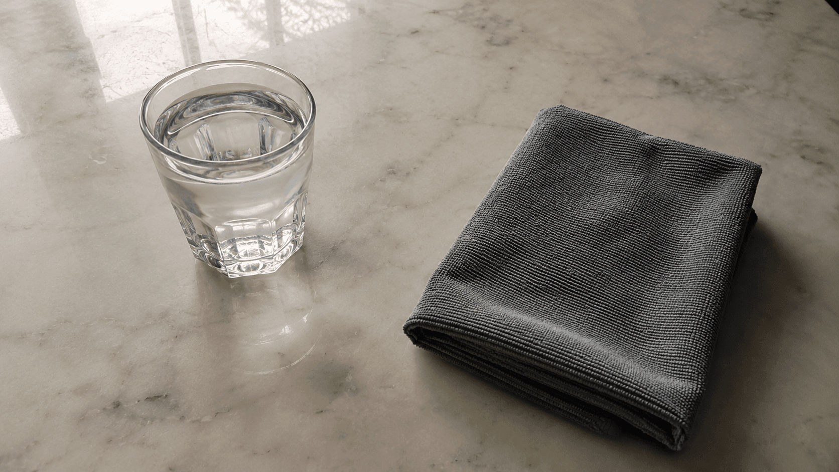 Minimal flat-lay: just a glass of plain water and one clean microfibre cloth on a marble surface — the only two things a coated shower needs